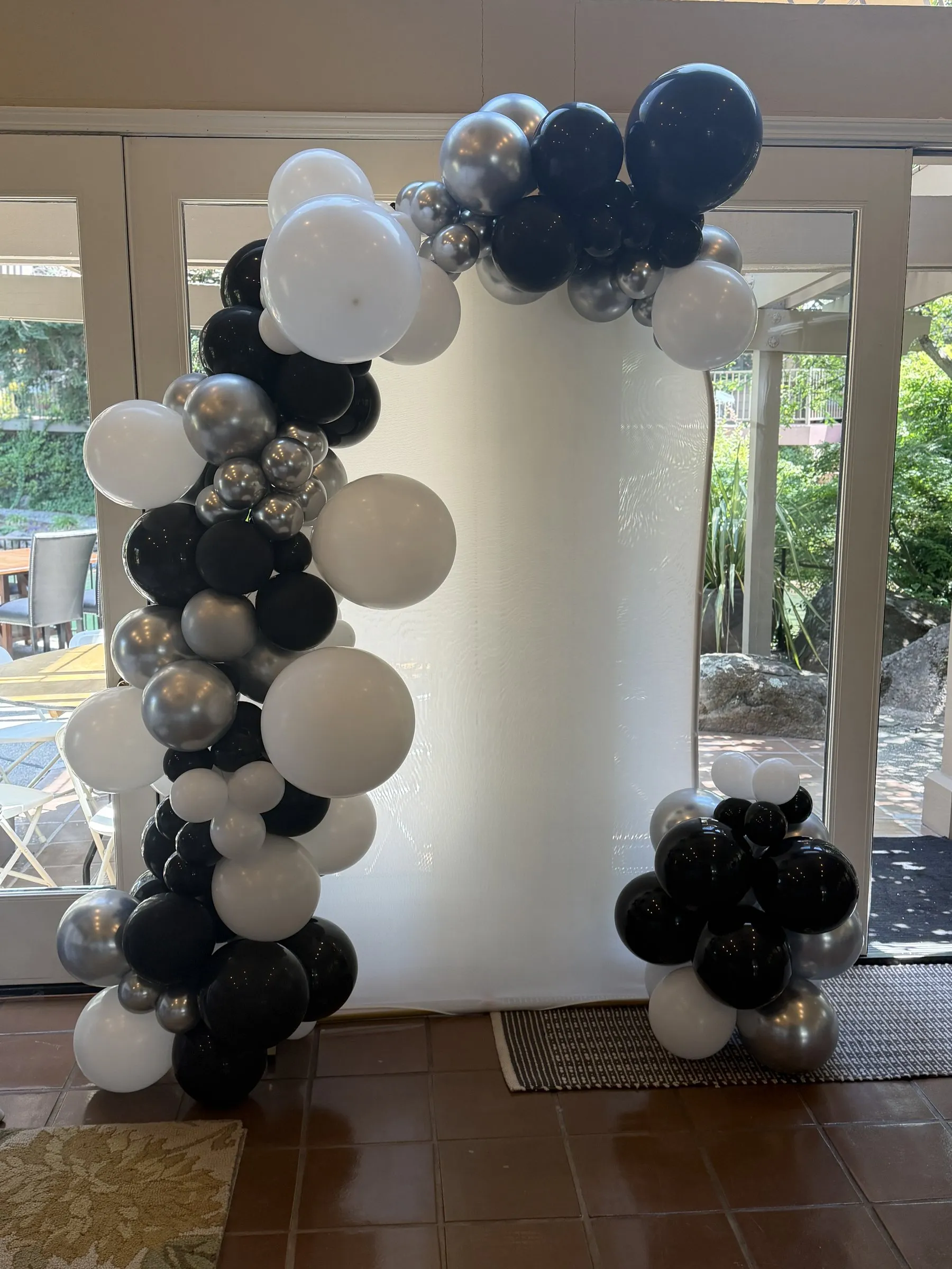 Black, white and chrome silver organic balloon arch styled around a tall white column backdrop in front of bright garden windows