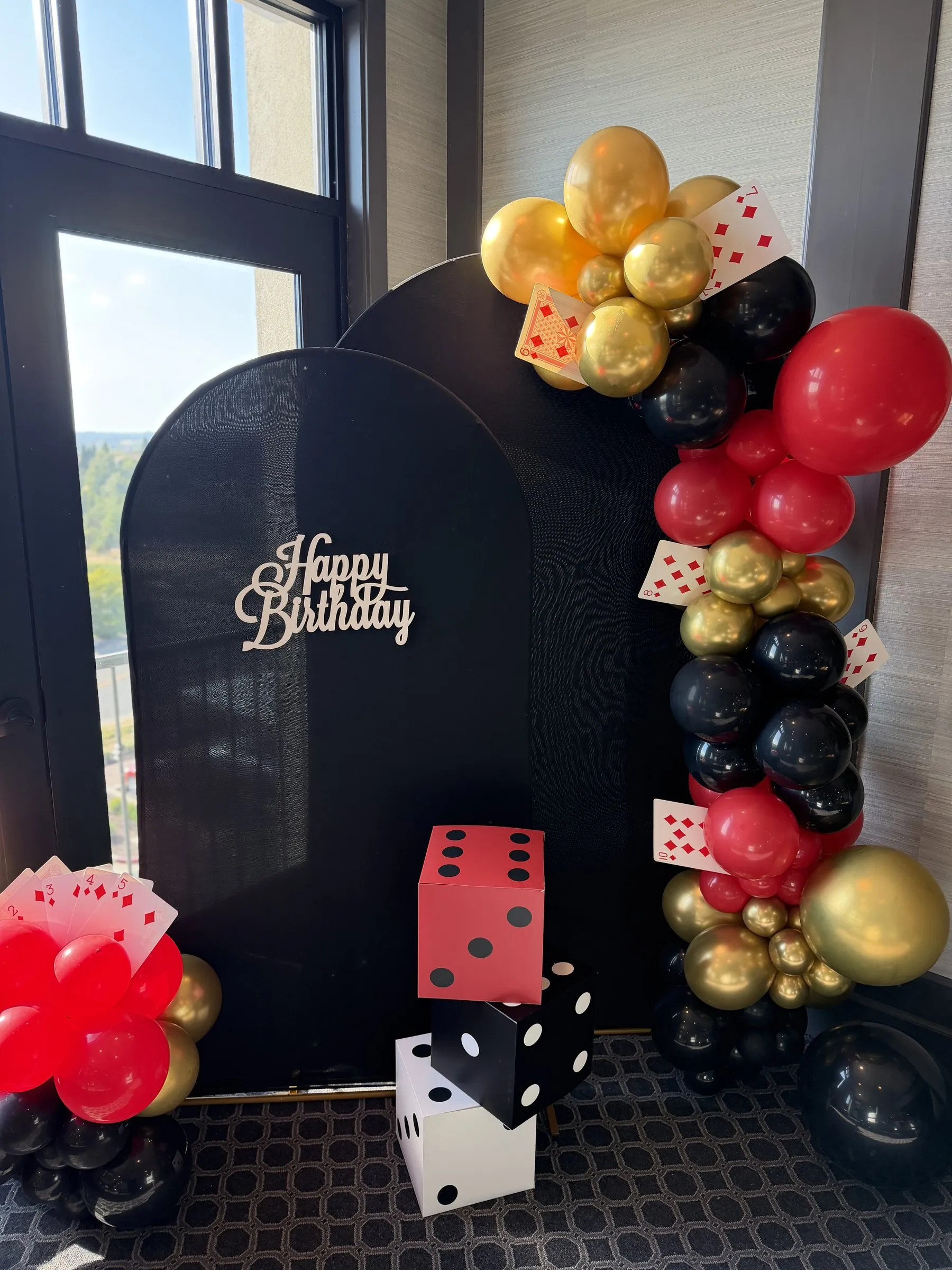 Full-height black balloon arch backdrop with a Happy Birthday script sign, red, black and gold garland and stacked dice props at a Folsom celebration