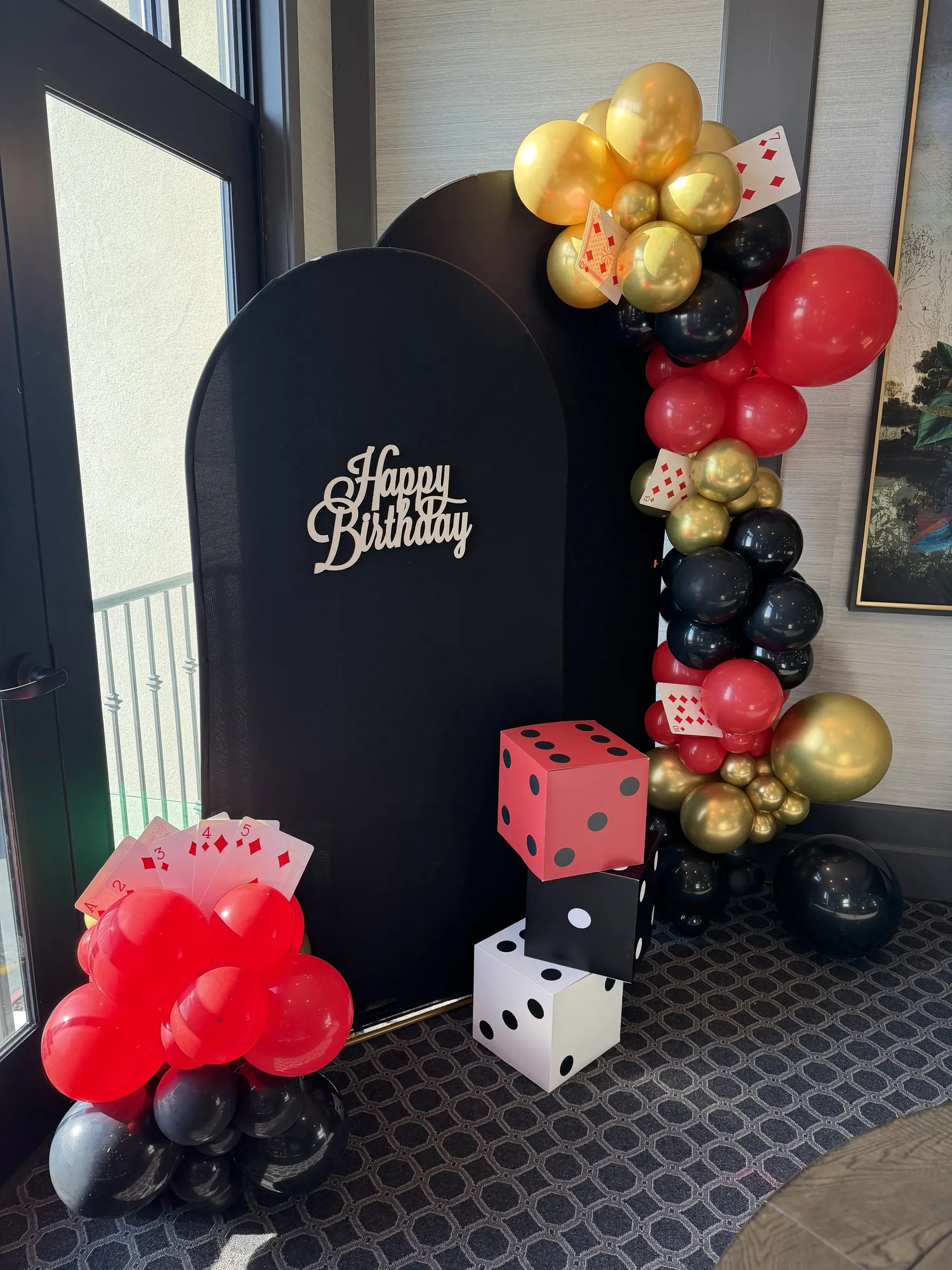 Red and black demi arch balloon cluster finished with a fanned playing-card accent beside a black arched backdrop