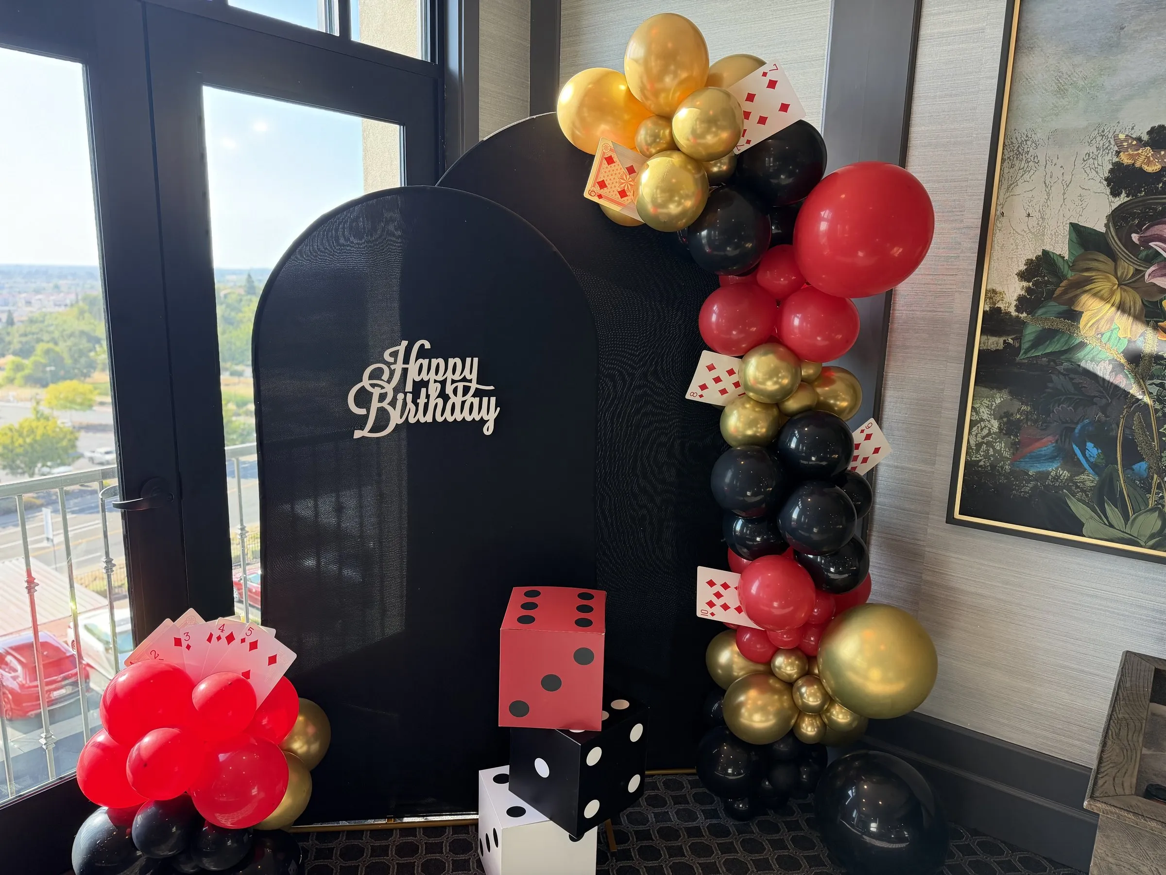 Casino-theme balloon backdrop styled in a clubhouse lounge — black arched panels with a red, black and gold organic garland, playing-card accents and oversized dice