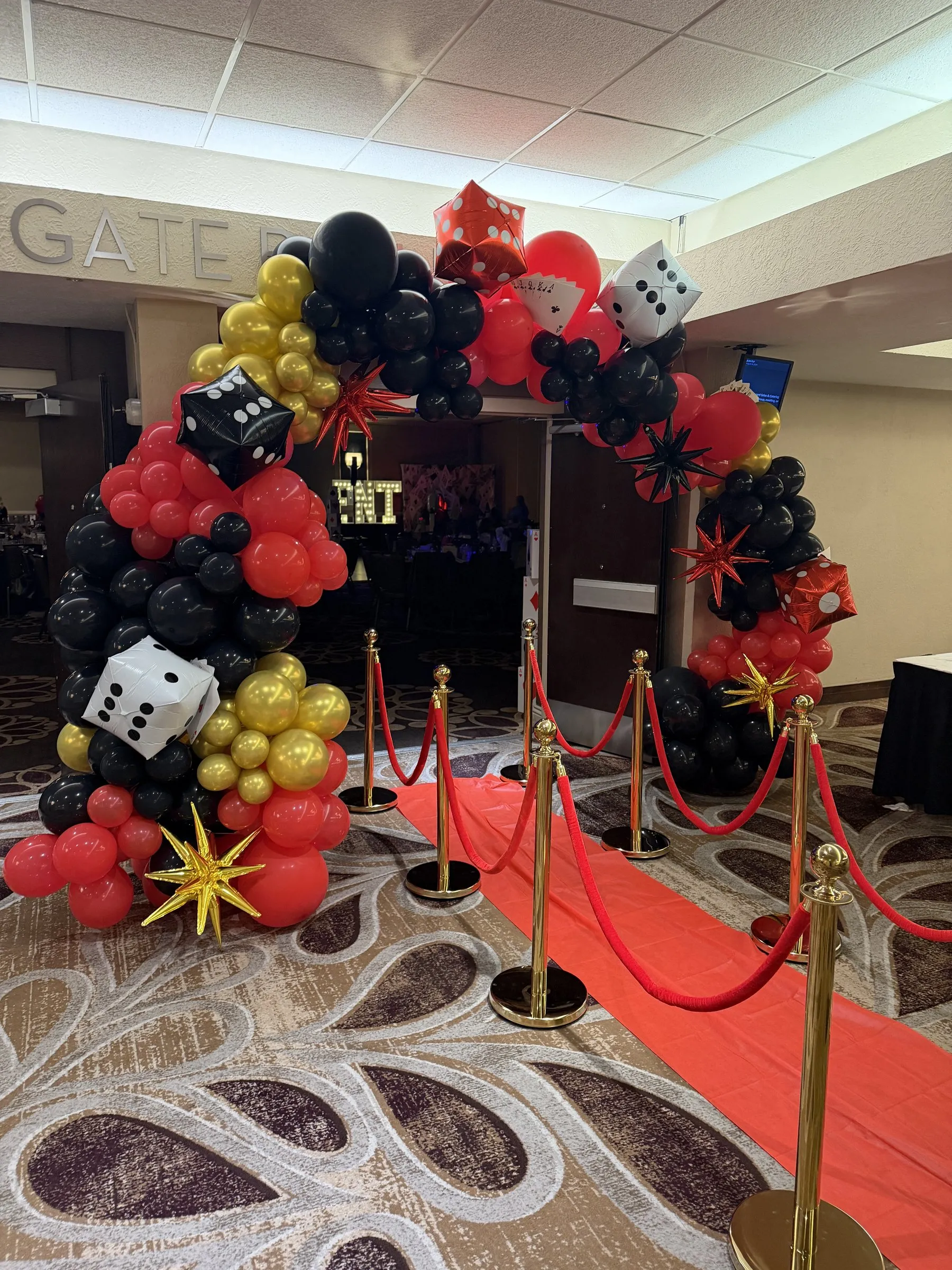 Red carpet entrance framed by a full walk-through balloon arch in red, black and gold with dice, playing cards and starburst accents at a corporate casino night