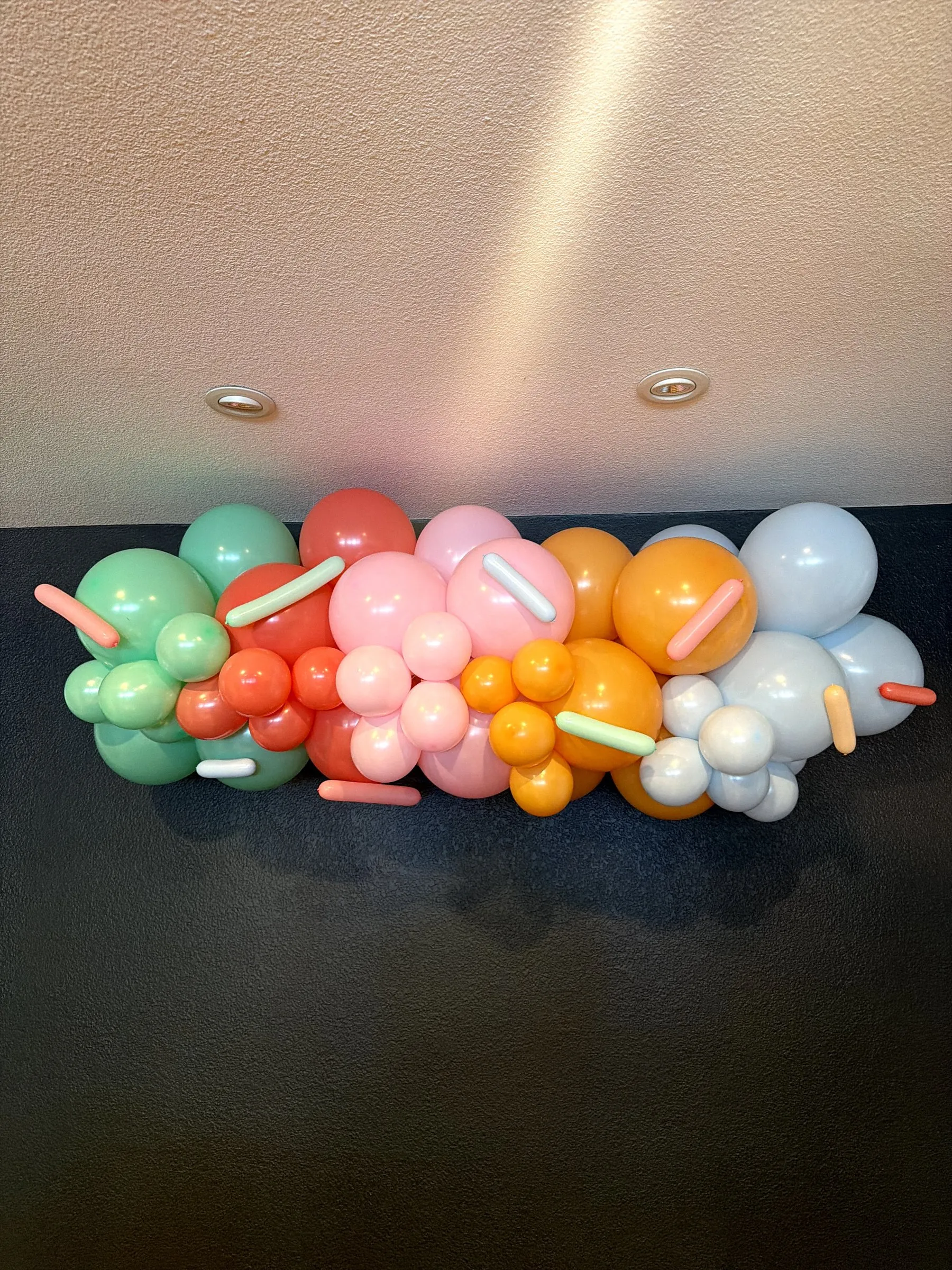 Donut-theme pastel balloon garland in mint, coral, pink, orange and powder blue installed on a dark accent wall for a children's birthday